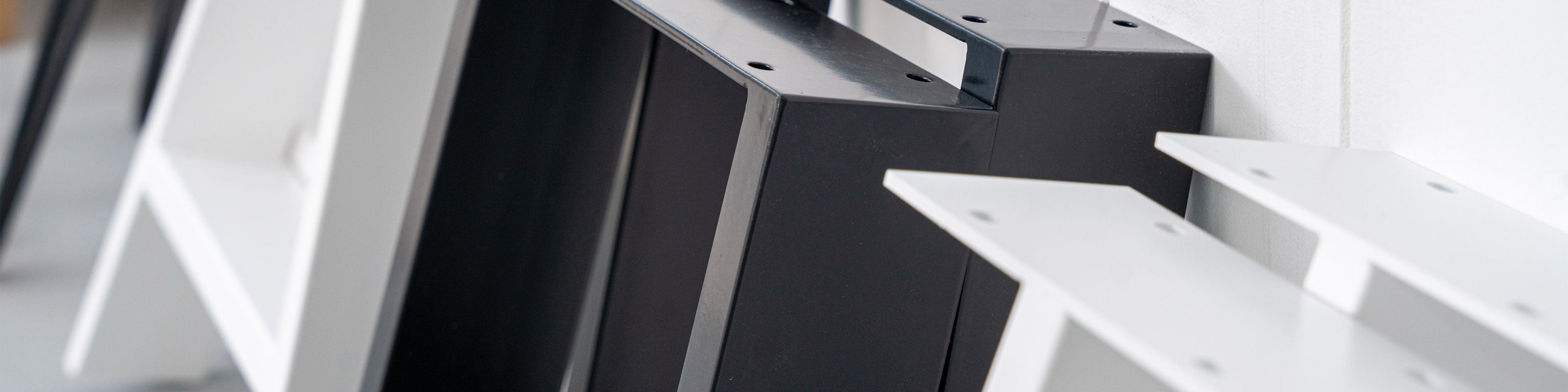 Black and white box-section table legs stood against a wall, top plates and fixing holes in view