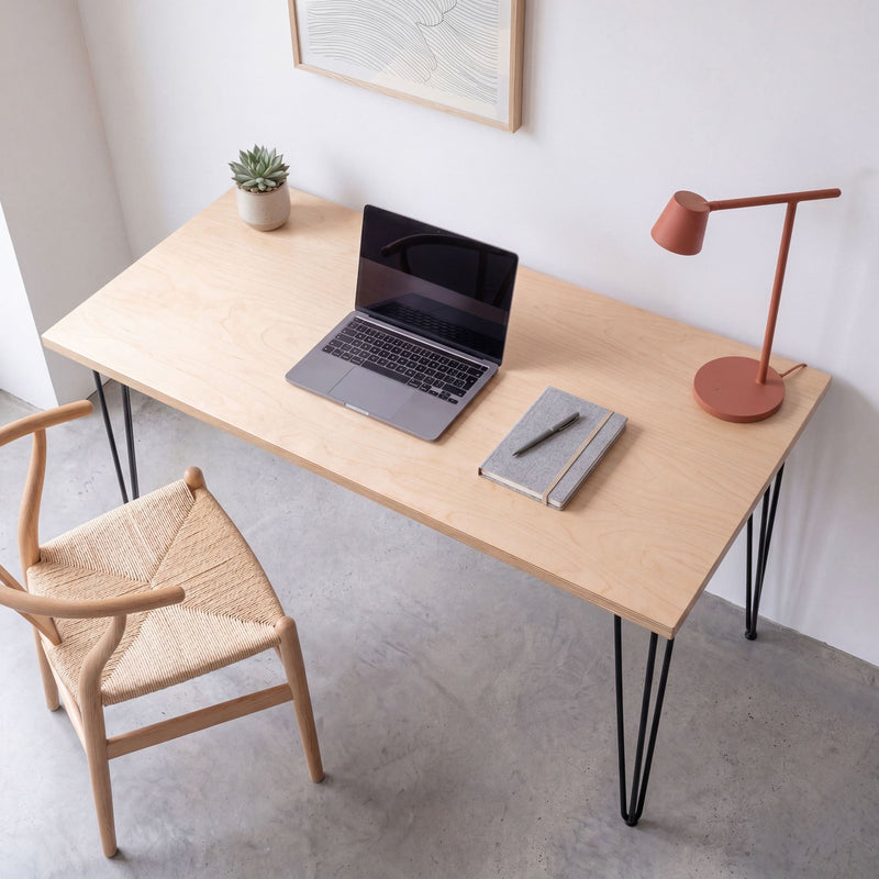 Angled view of a birch plywood desk with black hairpin legs, laptop and desk accessories